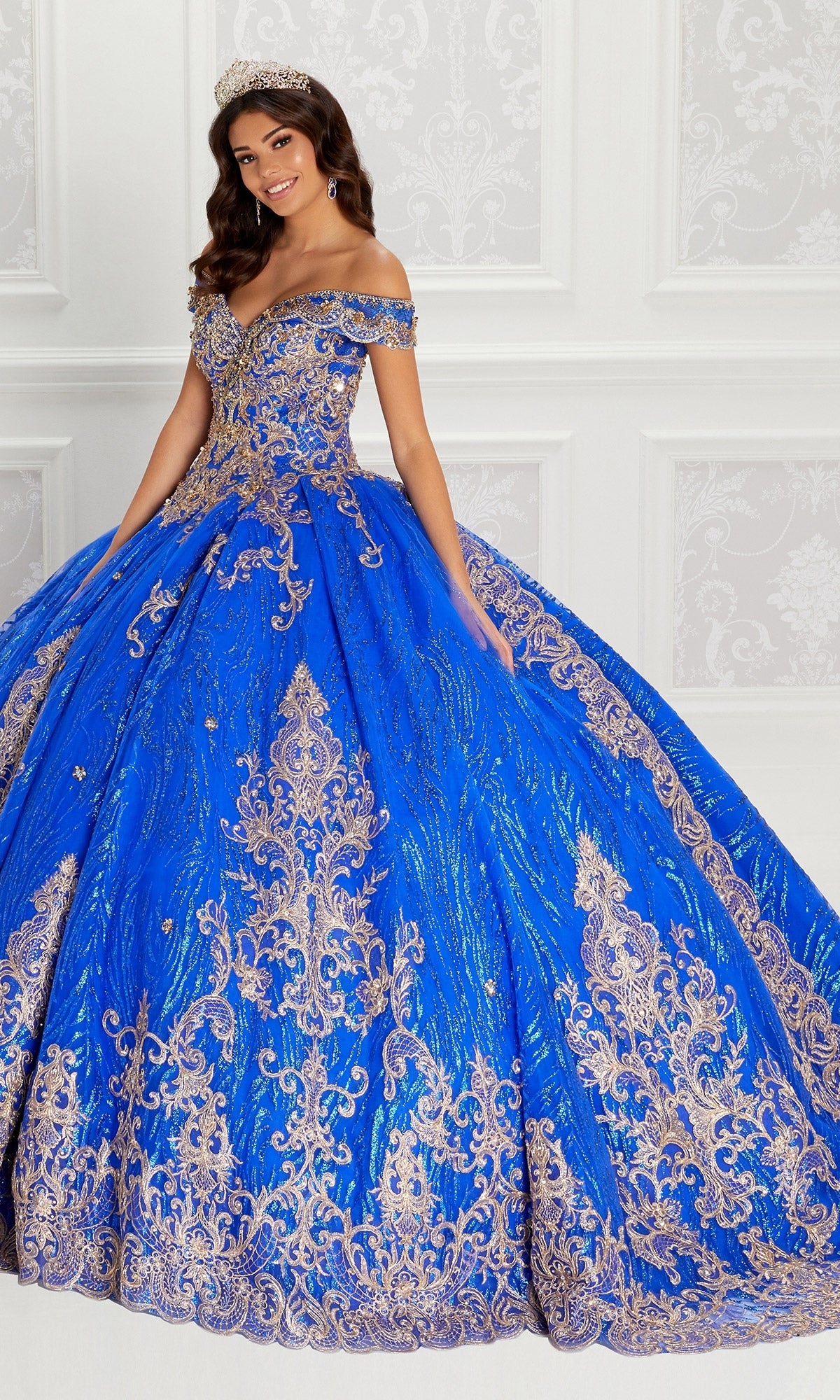 A woman poses against a white ornate wall wearing the bright blue Quinceanera Dress PR22148 By Princesa with gold lace embroidery and a matching tiara.